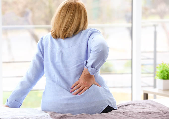 Woman sits on bed with hand on lower back to indicate back pain