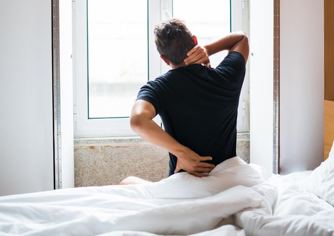 Man sits on edge of bed with hand on lower back indicating back pain from mattress